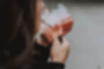A person holding a glass with a pink beverage and visible smoke or vapor rising from it, viewed from the side. The background is out of focus.