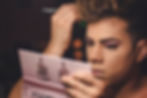 A person with short hair applies makeup to their face, holding a makeup palette in one hand and a brush in the other. The background is out of focus.