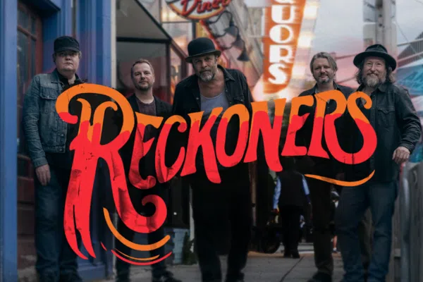 Five men in casual clothes stand on a city sidewalk with neon signs in the background. The word "Reckoners" in large, stylized red and orange text is overlaid across the center of the image.