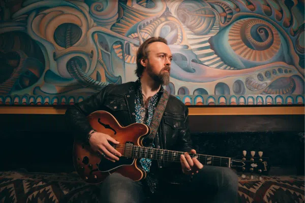 A man with a beard and mustache sits on a patterned sofa, playing an electric guitar. Wearing a leather jacket and patterned shirt, he channels the vibe of the Gravel Project. Behind him is a large, colorful abstract mural with swirling shapes.