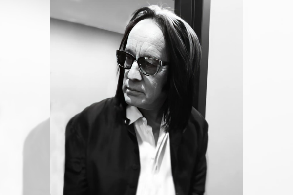 A man with shoulder-length dark hair and white streaks wears sunglasses, a white shirt, and a dark jacket, looking to the side in a well-lit indoor setting.