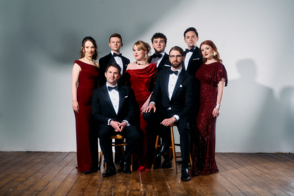 A group of four men in tuxedos and four women in red formal dresses pose together indoors against a plain white wall and wooden floor; two men are seated while the others stand.