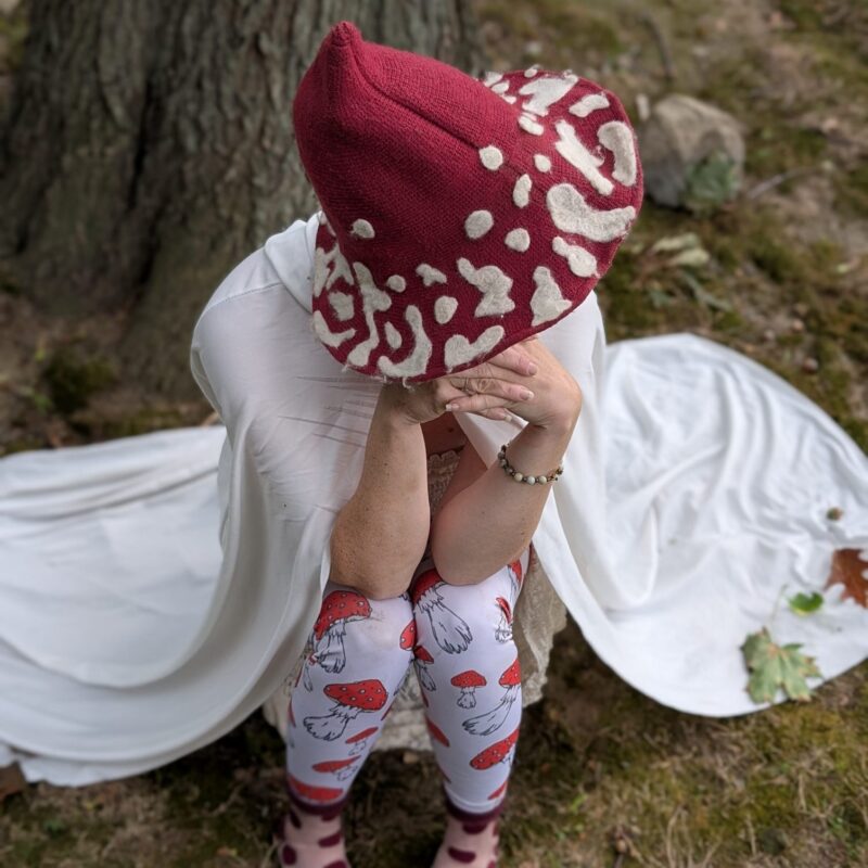 A person sits on grass near a tree, wearing a red and white mushroom-patterned hat, a white cape, and leggings with red mushrooms. Their face is covered with their hands.