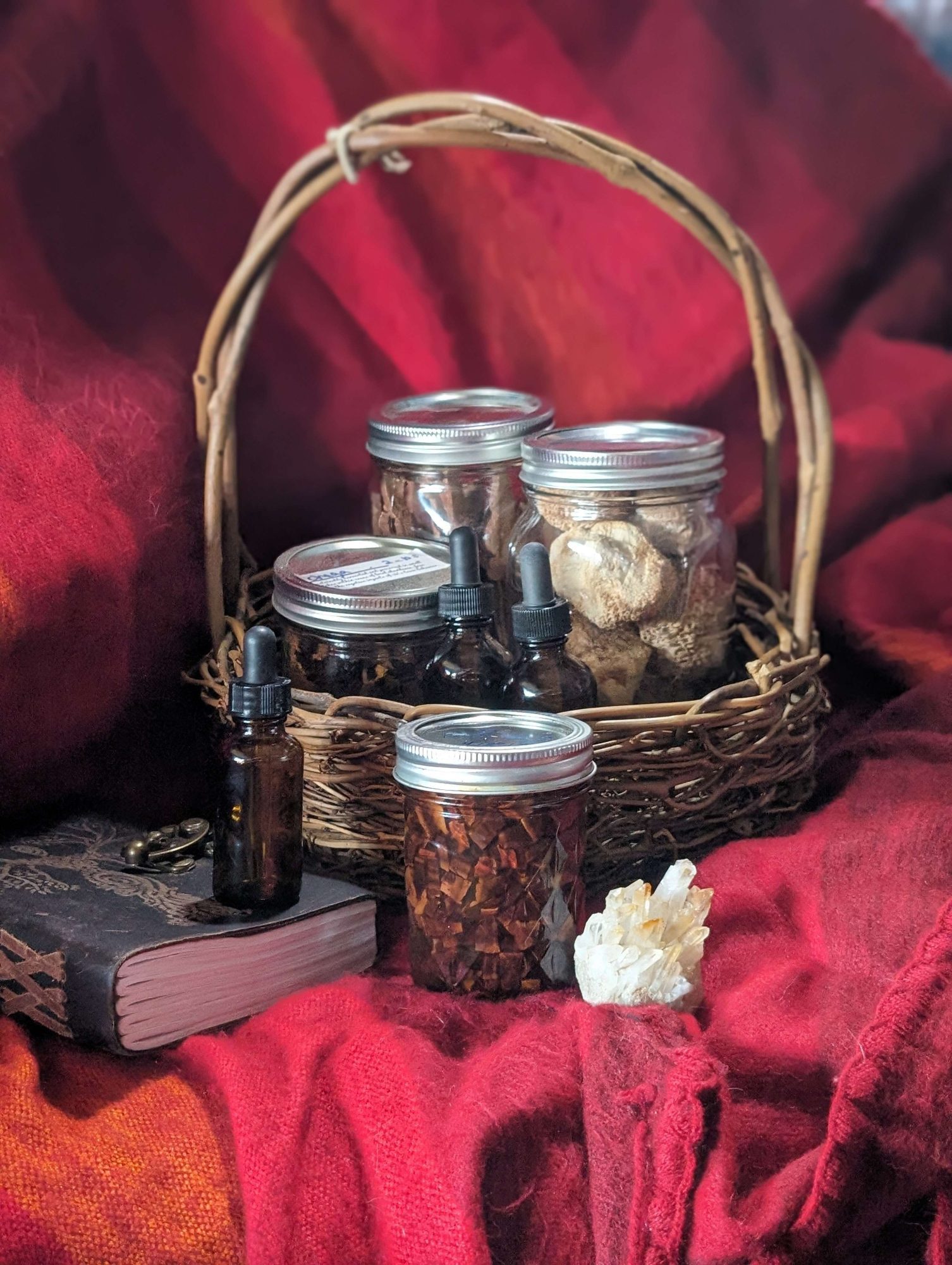 A wicker basket on a red cloth holds jars of dried herbs or mushrooms and two dropper bottles. In front are a large crystal, a brown jar with dried herbs, and a closed, ornate book with a key.