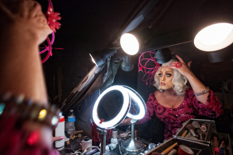 A person wearing a sequined pink outfit and a white wig looks into a lit vanity mirror while attaching a pink flower accessory to their hair, surrounded by makeup and hairstyling tools.