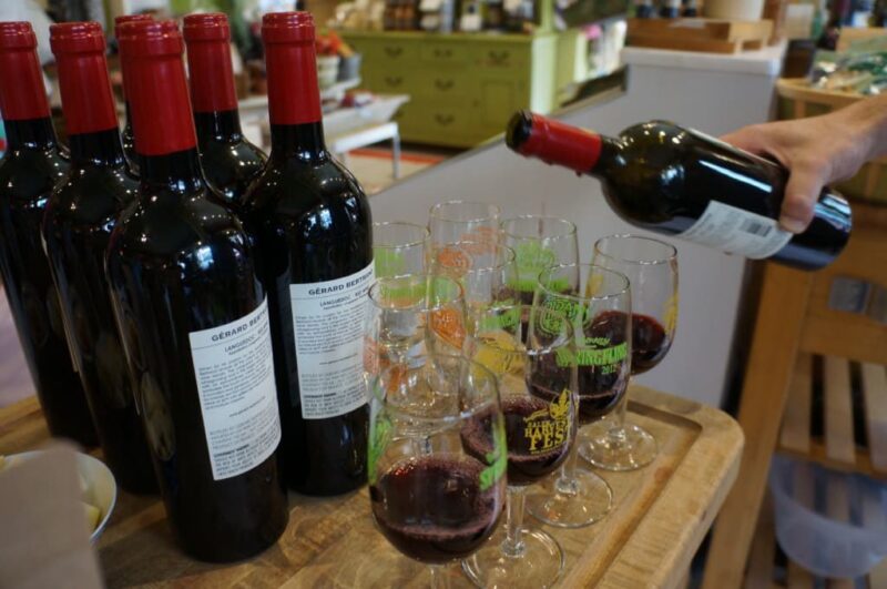 Bottles of red wine are lined up next to several glasses, some of which are being filled with red wine by a hand holding a bottle. The scene appears to be set on a wooden surface in a store or tasting area.