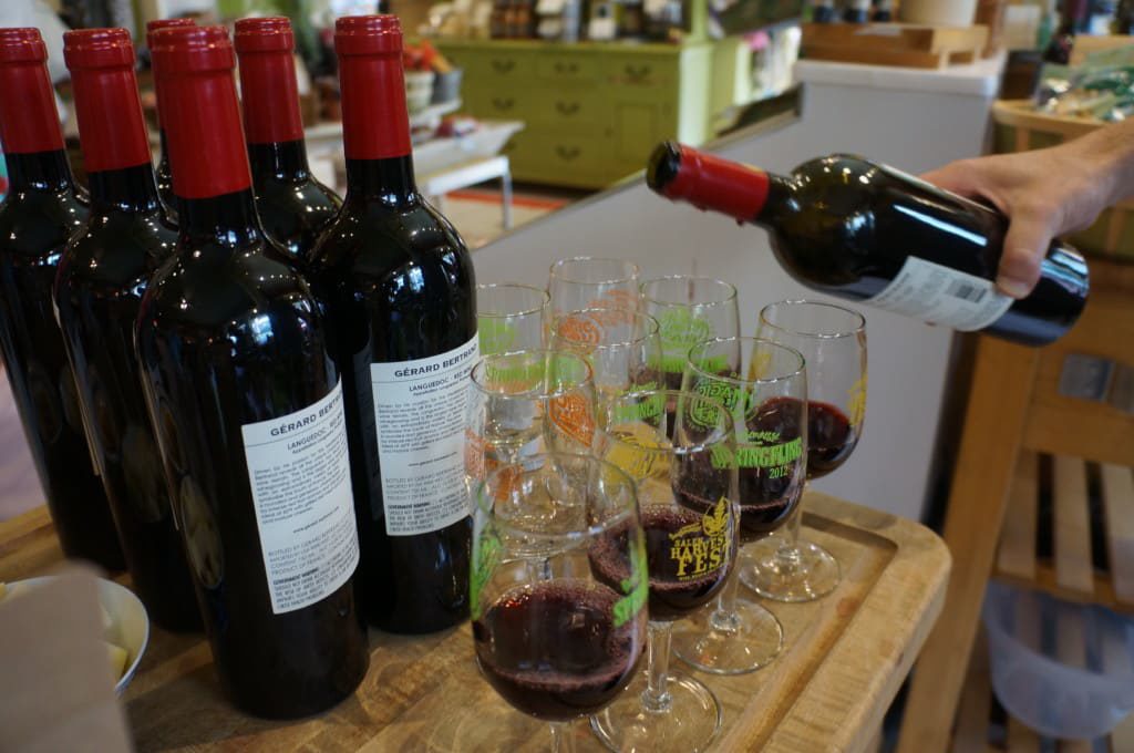 Bottles of red wine are lined up next to several glasses, some of which are being filled with red wine by a hand holding a bottle. The scene appears to be set on a wooden surface in a store or tasting area.