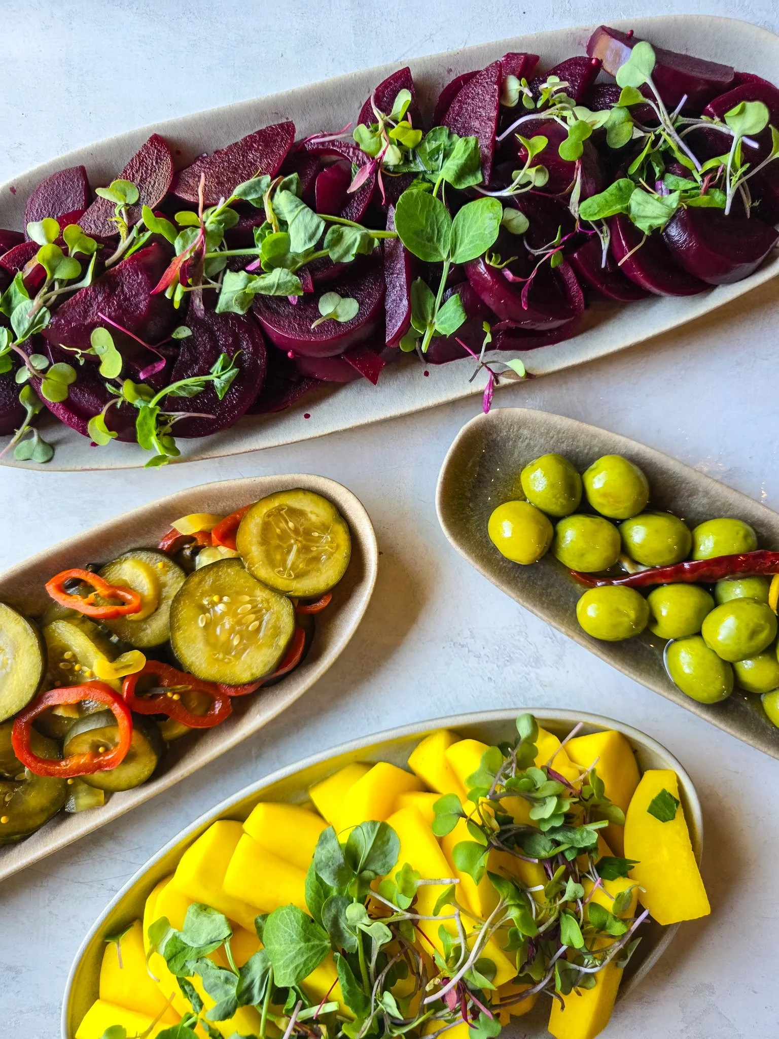 Four serving dishes with assorted appetizers—sliced beets with microgreens, green olives, pickled cucumbers with red peppers, and yellow pickled vegetables—beautifully arranged on a light surface for Sevenings at CCE Nightlife in Essex County.