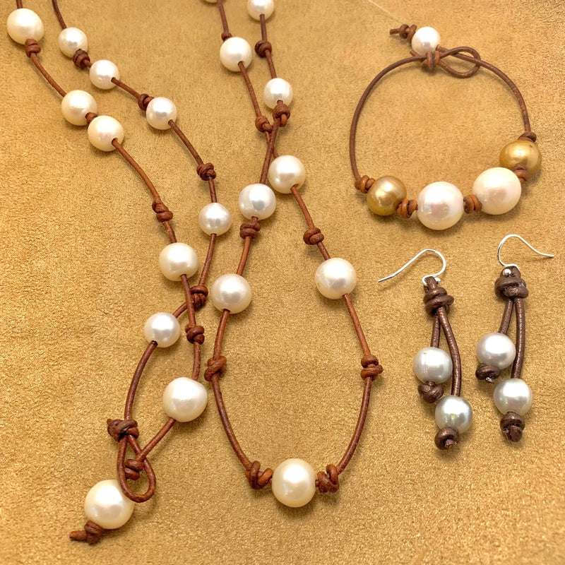 A necklace, bracelet, and earrings made of brown cord and white, gold, and silver pearls are displayed on a tan surface. The necklace and bracelet feature scattered pearls, while the earrings have two pearls each.