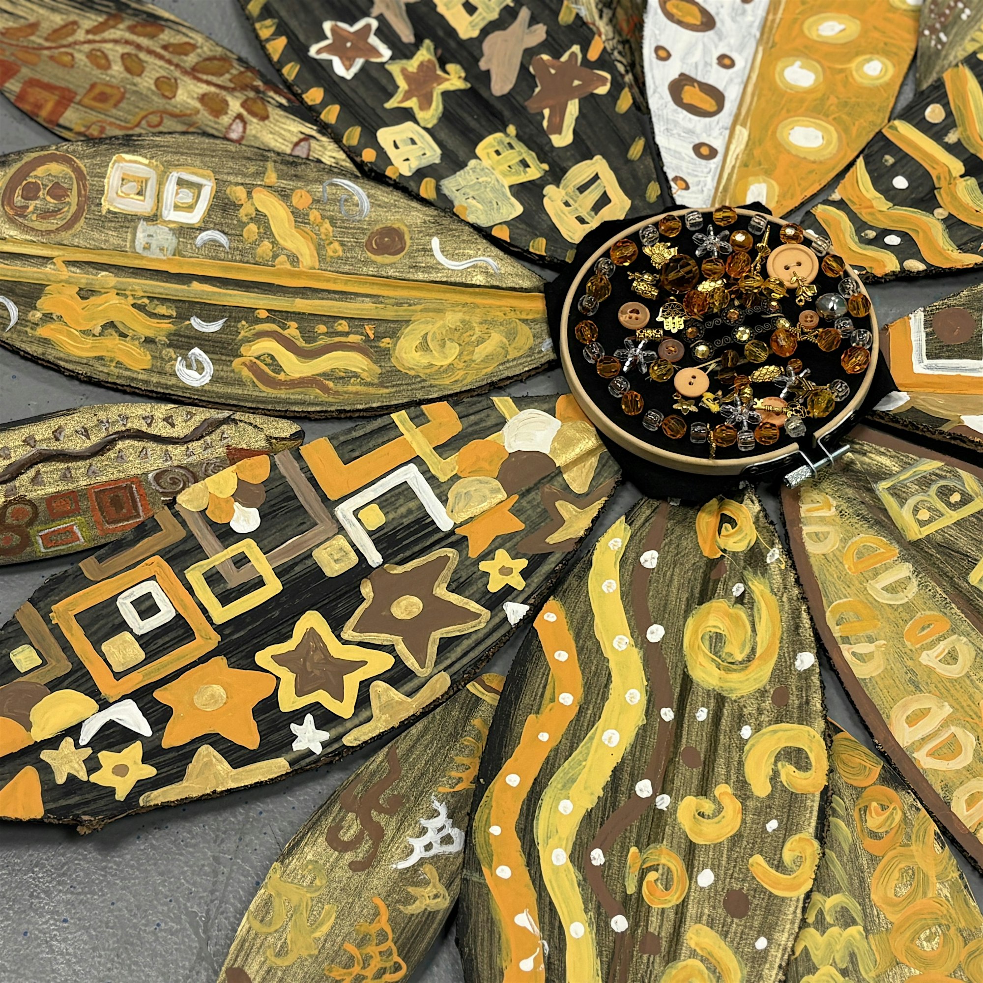 A close-up of a decorative flower made from painted, patterned petals in yellow, gold, brown, and black. The petals feature geometric and swirl designs. The center is filled with beads and buttons in similar colors. The background is gray.