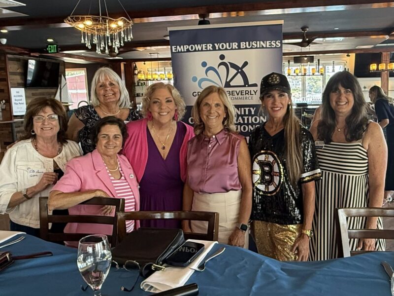 Seven women stand together in a restaurant, posing for a group photo in front of a "Greater Beverly Chamber of Commerce" banner. The table in front has blue tablecloths and glassware. The atmosphere appears friendly and professional.