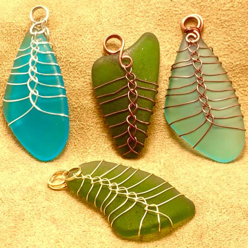 Four sea glass pendants, two blue and two green, each wrapped in wire with leaf and web-like patterns, are displayed on a light tan fabric background.