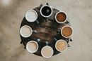 Eight cups with different shades of coffee ranging from light to dark are arranged in a circle on a wooden table, viewed from above. The table surface is rustic, and the background is a neutral, blurred floor.