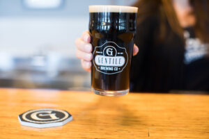 A person holds a pint glass filled with dark beer from Gentile Brewing Co. on a wooden counter, perfect for enjoying Sevenings and exploring Essex County nightlife. A matching coaster sits in the foreground, while the background is softly blurred.