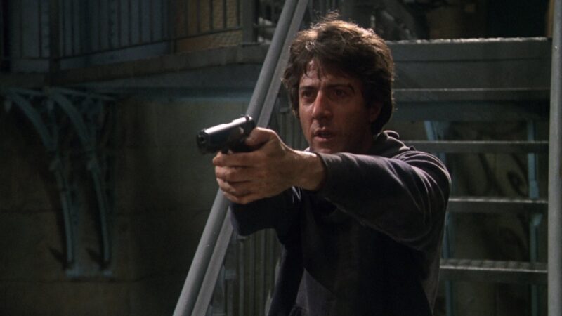 A man with tousled dark hair, wearing a dark sweatshirt, stands indoors aiming a handgun with both hands. Metal stairs and railings are visible in the background.