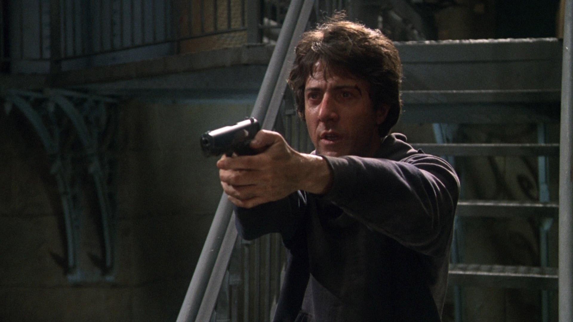 A man with tousled dark hair, wearing a dark sweatshirt, stands indoors aiming a handgun with both hands. Metal stairs and railings are visible in the background.