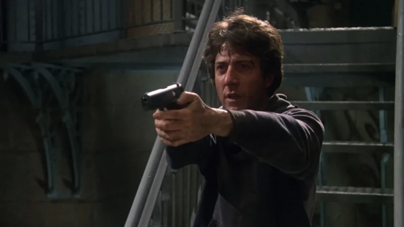 A man with dark hair points a handgun forward with both hands, standing near a metal staircase in a dimly lit, industrial-looking setting—a tense moment captured from the Marathon Man 50th Anniversary Series.