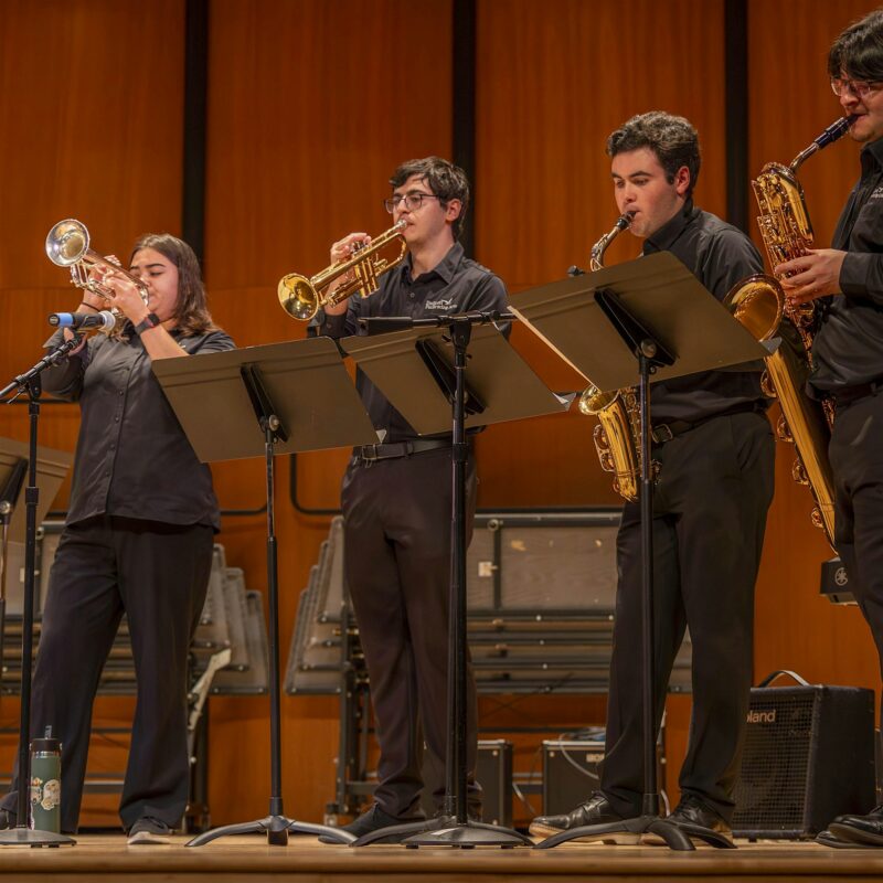 Four musicians dressed in black perform on stage, each playing a brass instrument. They stand behind music stands with sheet music, and a wooden backdrop and music equipment are visible behind them.