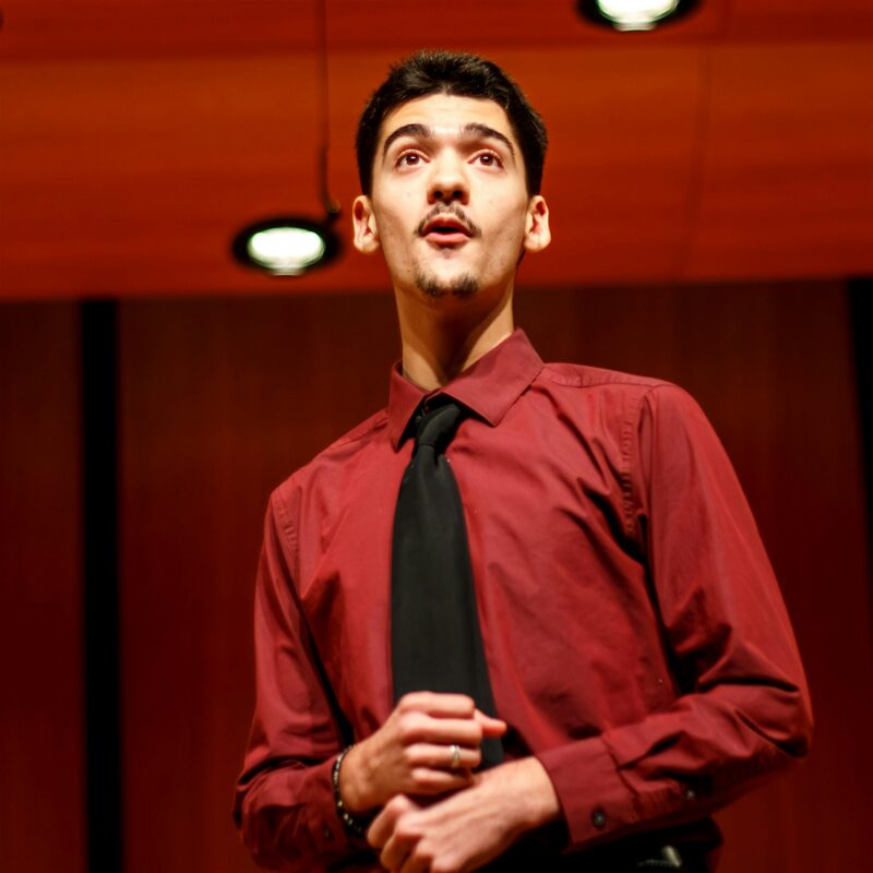 A young man wearing a maroon shirt and black tie stands under stage lights, looking slightly upwards with a focused expression. He holds his hands together in front of his chest. The background shows a wooden-paneled wall and ceiling lights.