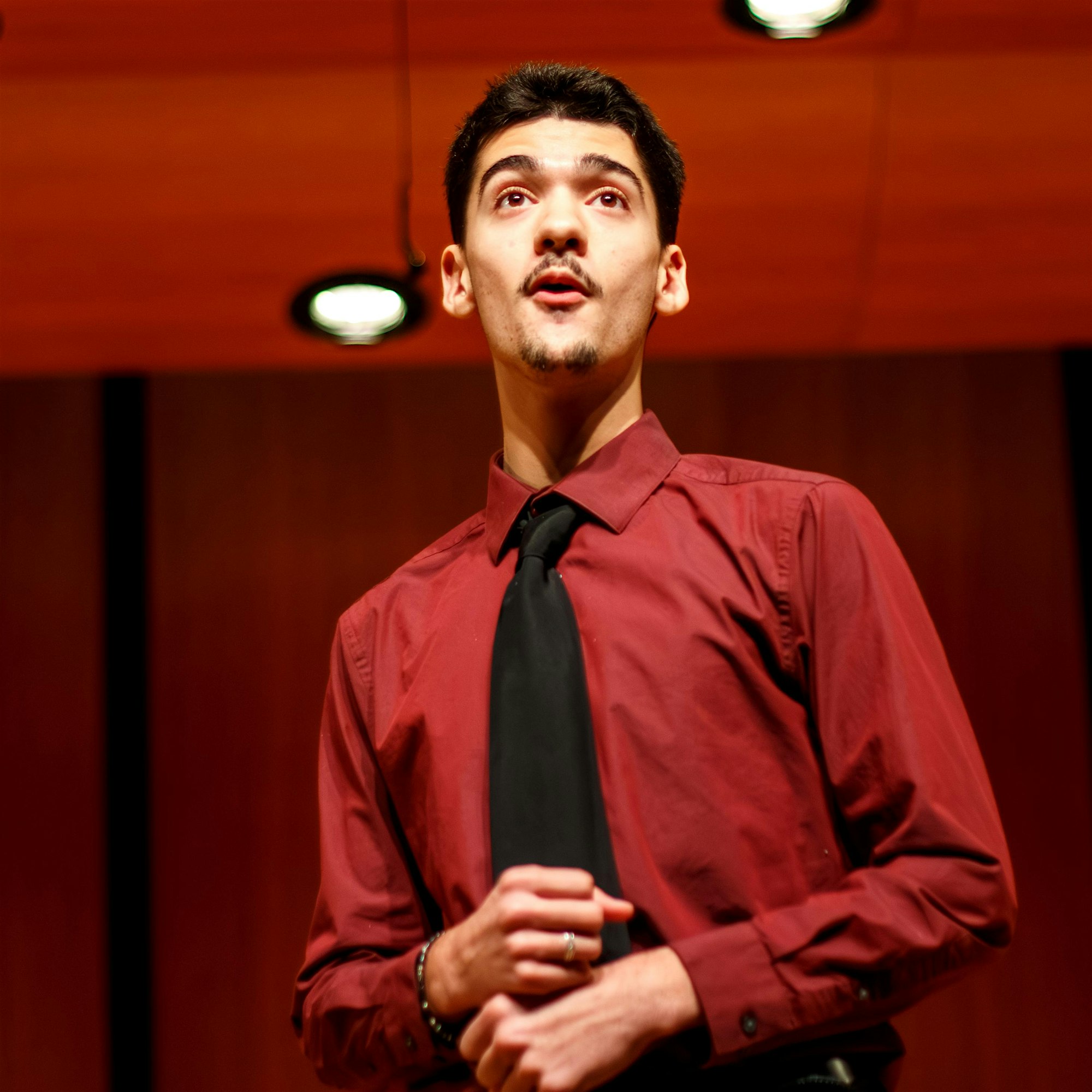 A young man wearing a maroon shirt and black tie stands under stage lights, looking slightly upwards with a focused expression. He holds his hands together in front of his chest. The background shows a wooden-paneled wall and ceiling lights.