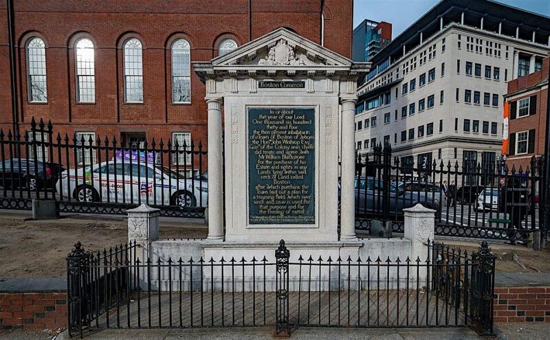 A stone monument with an inscribed plaque stands behind a black iron fence. The monument is situated on a paved area, with a brick building and parked cars visible in the background.