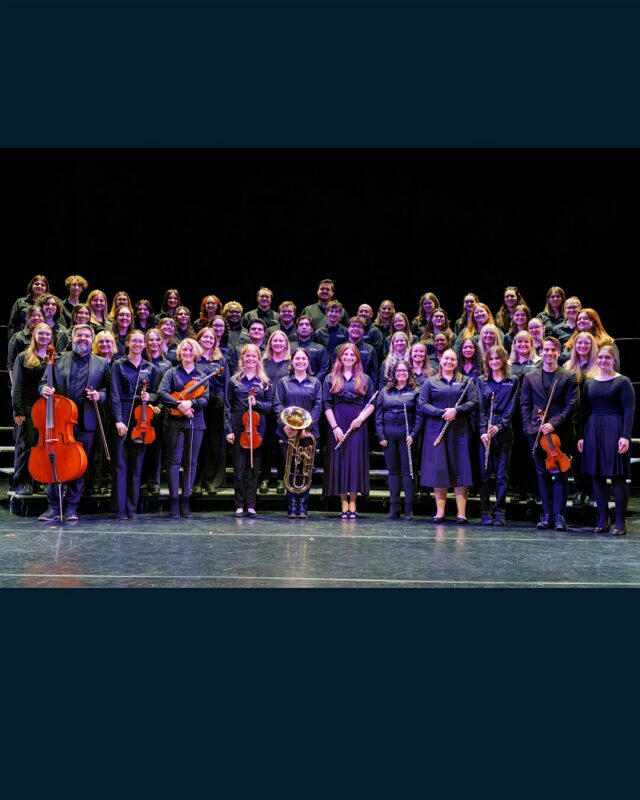 A large group of musicians, dressed in black, stand on stage with their instruments, including strings and brass, arranged in rows and smiling at the camera.
