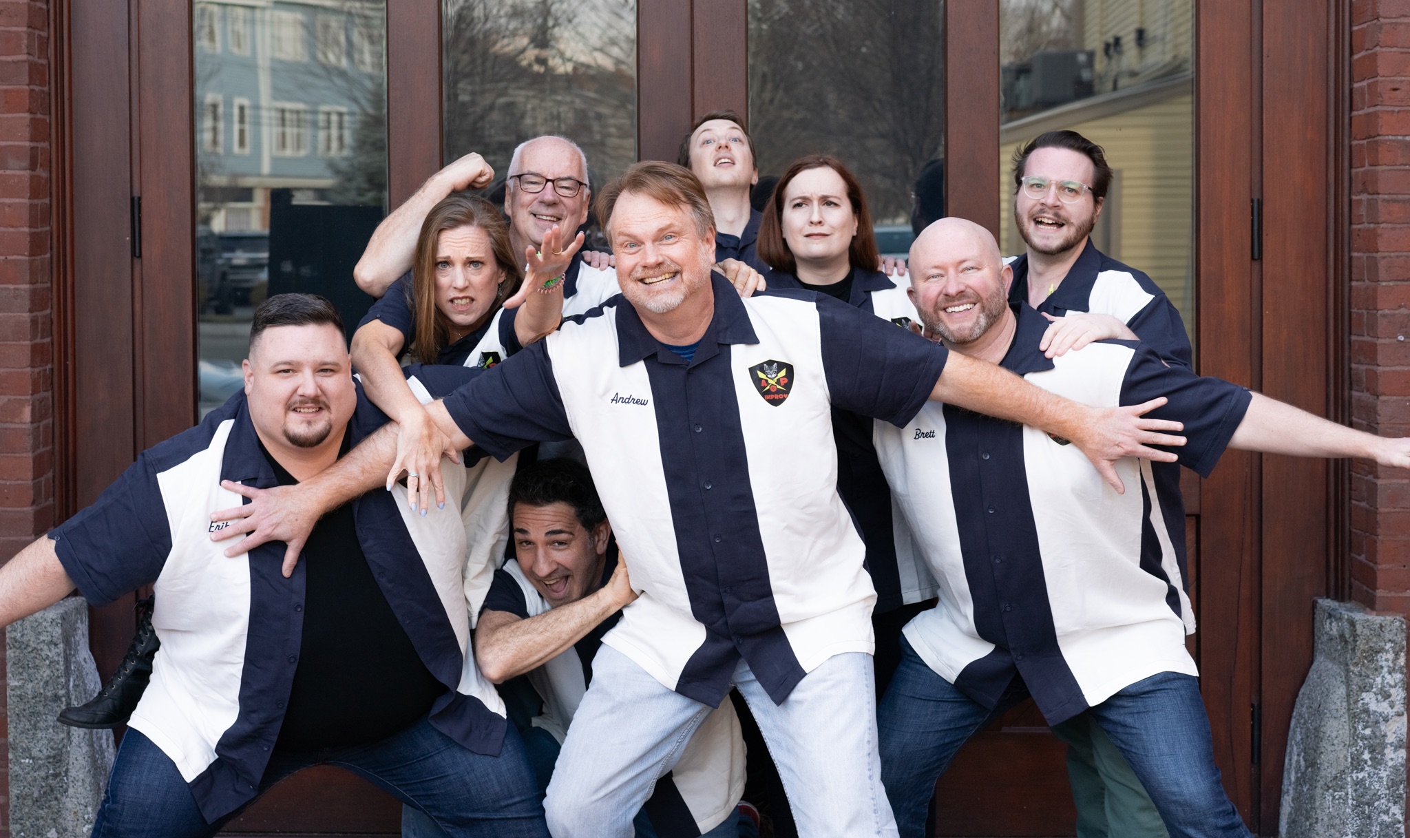 Eight adults in matching bowling shirts pose playfully by glass doors—some smiling, some making enthusiastic gestures, channeling the spirit of improv comedy in Marblehead as one stands prominently in front, others grouped closely behind and to the sides.