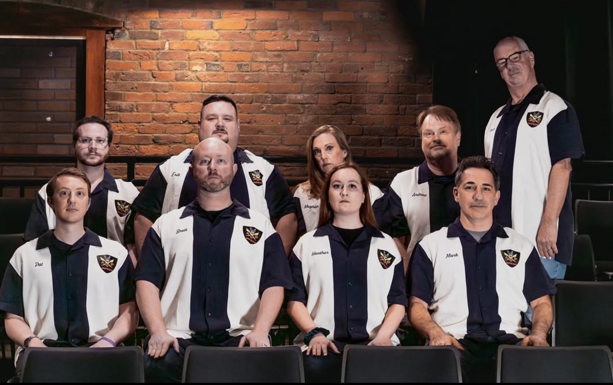 Nine adults wearing matching bowling-style shirts with embroidered patches pose for a group photo in front of a brick wall—cast members after an improv comedy in Marblehead. Four are seated on theater-style black chairs, while five stand behind them indoors.