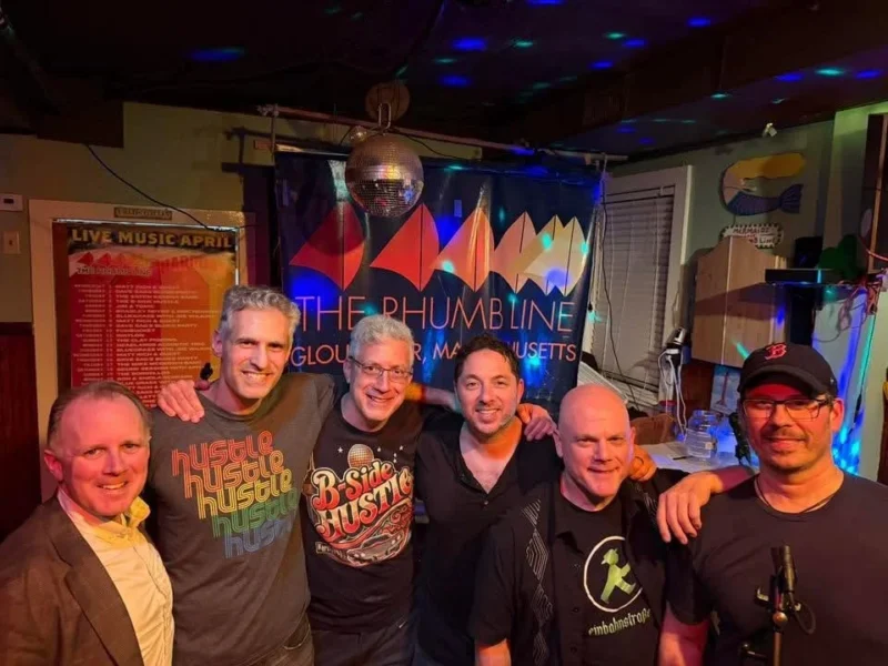 Six men stand together smiling in front of a banner reading "The Phumbl Line" with colorful graphics. A disco ball hangs above them, and there are spotlights and posters visible in the background. Some are wearing t-shirts with printed designs.