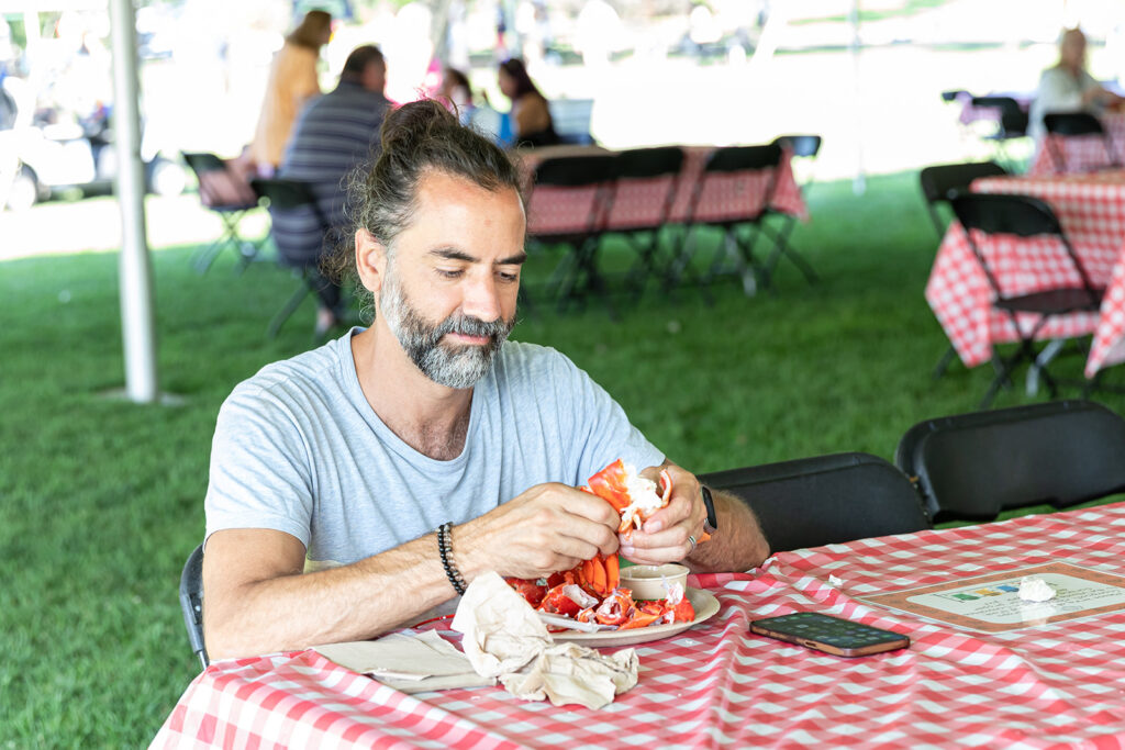 At a Beverly Homecoming 2026 event, a gray-haired man with a beard sits alone at a table with a checkered cloth, eating lobster outdoors. Nearby are empty chairs, other diners, and on his table—his phone, napkins, and menu.
