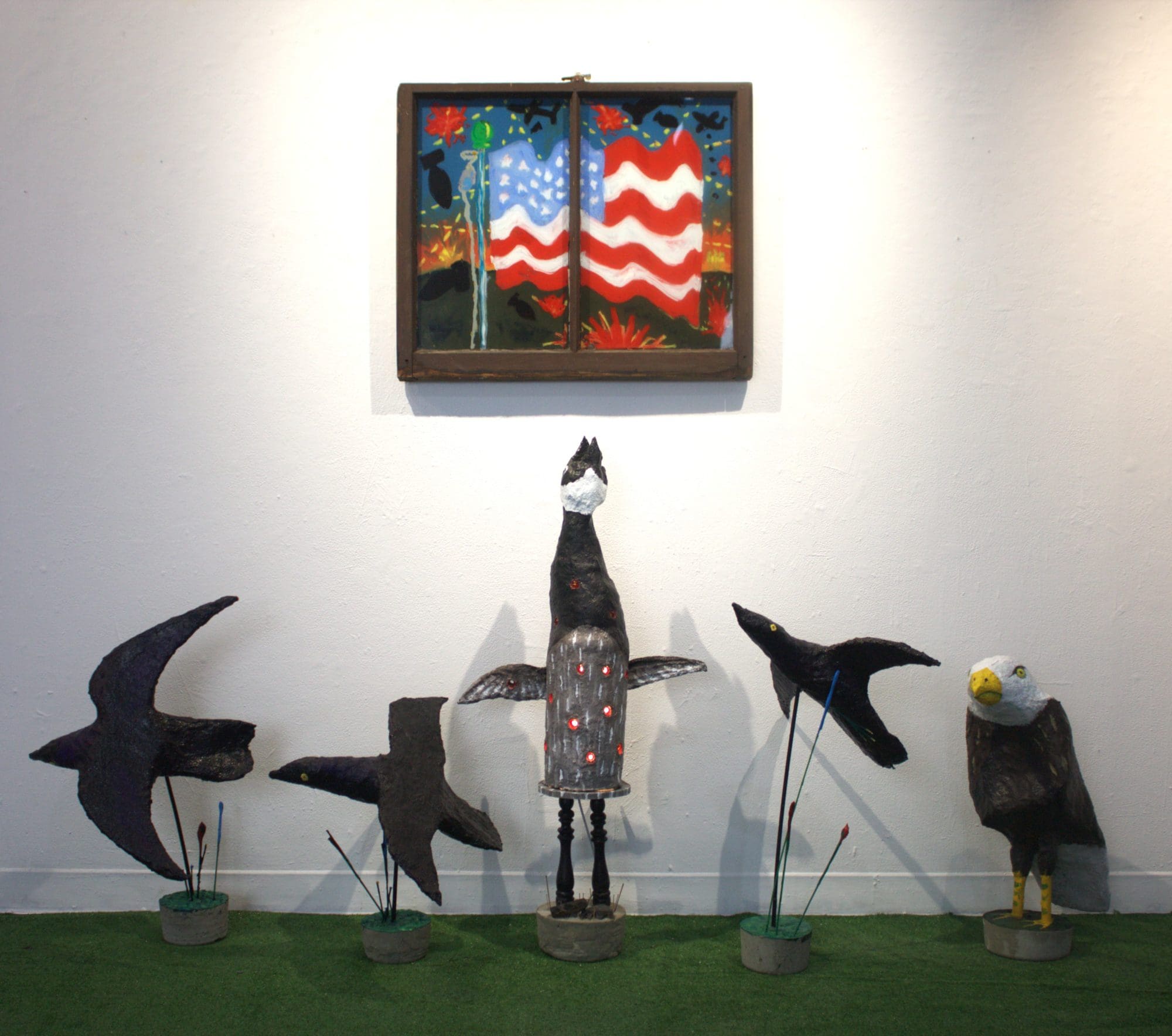 A painting of two wavy American flags hangs above five bird sculptures on a patch of artificial grass; three birds are black, one is black with lights, and one is an eagle with a yellow beak looking to the side.