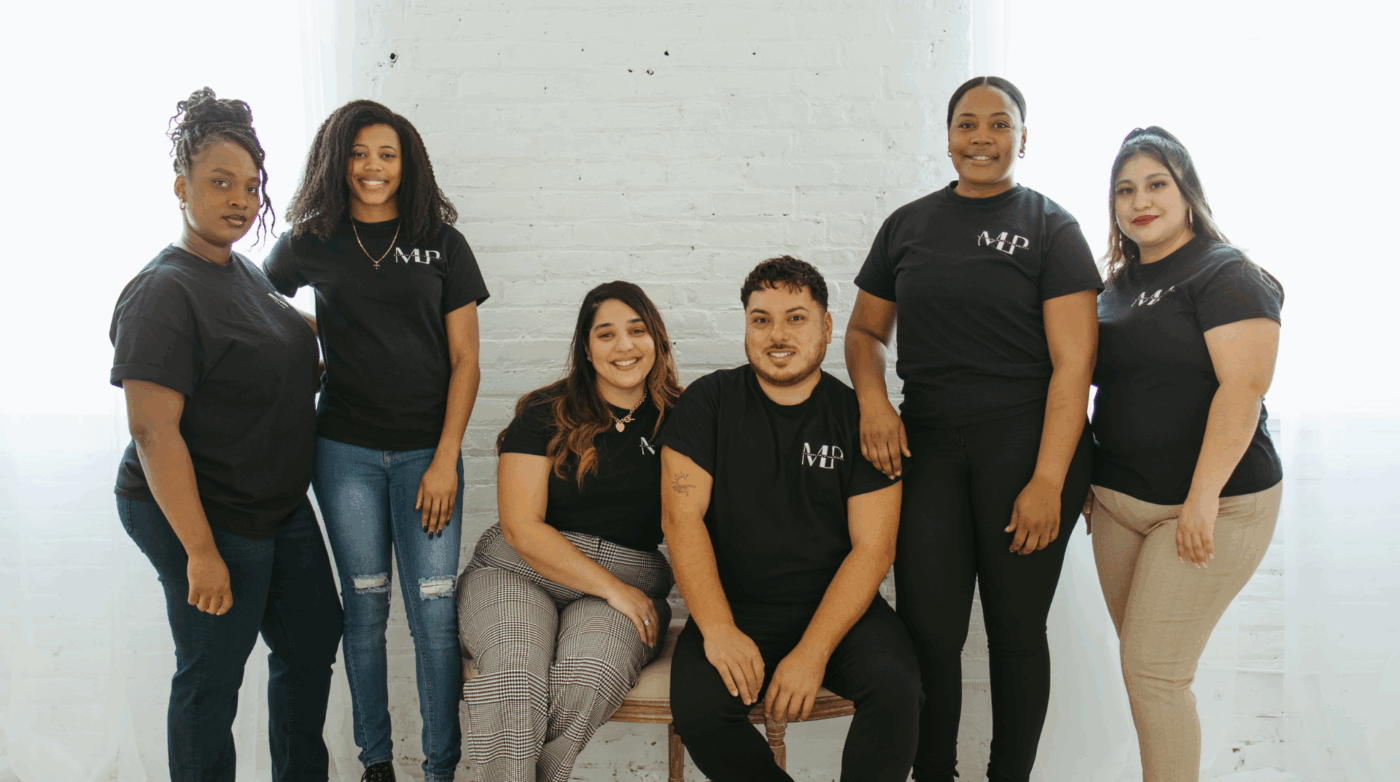 Six people pose indoors against a white brick wall, all wearing black t-shirts with a white logo and casual pants. The group is smiling at the camera, highlighting unity and support for Minority Mental Health Awareness.