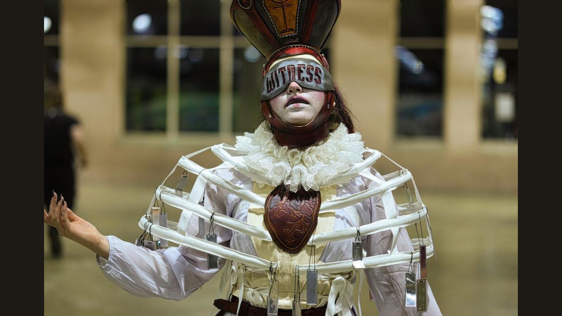 Meet the Member: Change is Simple—a person in an elaborate costume with a heart-shaped headpiece, “WITNESS” over their eyes, ruffled collar, white face paint, and a unique cage-like torso stands indoors with one arm slightly raised.