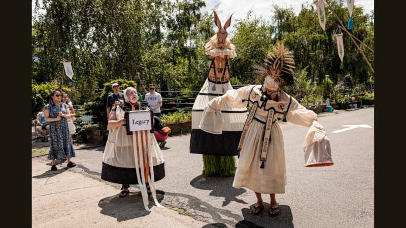 Two people in cream-colored costumes, including Alisa Sickora, stand on a street—one holds a “Legacy” sign. A large, horse-like puppet and decorated banners appear as several onlookers watch amid the surrounding trees.