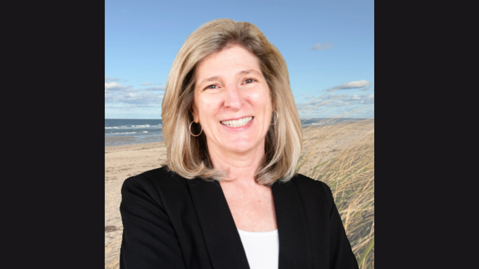 Meet the Member: MVP Arts—a woman with shoulder-length blonde hair, in a black blazer and white top, stands outdoors before a sandy beach, grassy dunes, ocean waves, and a blue sky dotted with clouds.
