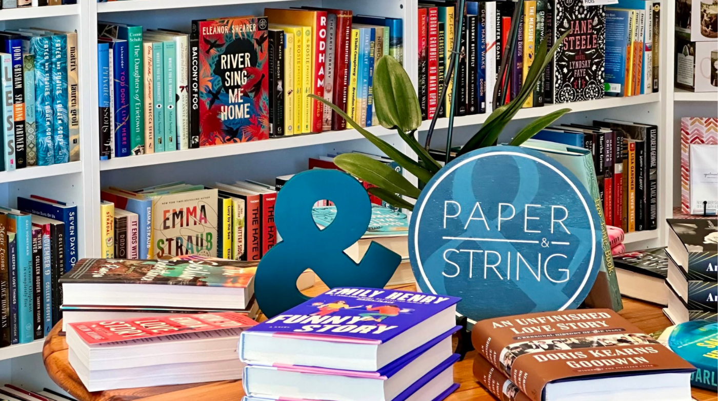 A bookstore display features stacks of books on a table, a small plant, a blue ampersand, and a round "Paper & String" sign, with colorful books and shelves creating a vibrant backdrop.