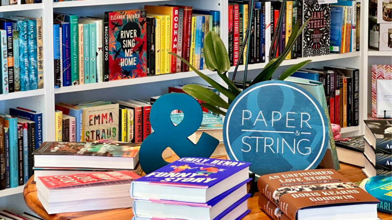 A bookstore display features stacks of books on a table, a small plant, a blue ampersand, and a round "Paper & String" sign, with colorful books and shelves creating a vibrant backdrop.
