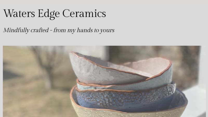 Four handmade Waters Edge Ceramics bowls, each with a unique shape and texture, are stacked outdoors. The top bowl is light pink and gray, followed by pale pink, textured blue, and textured tan. Background is blurred.