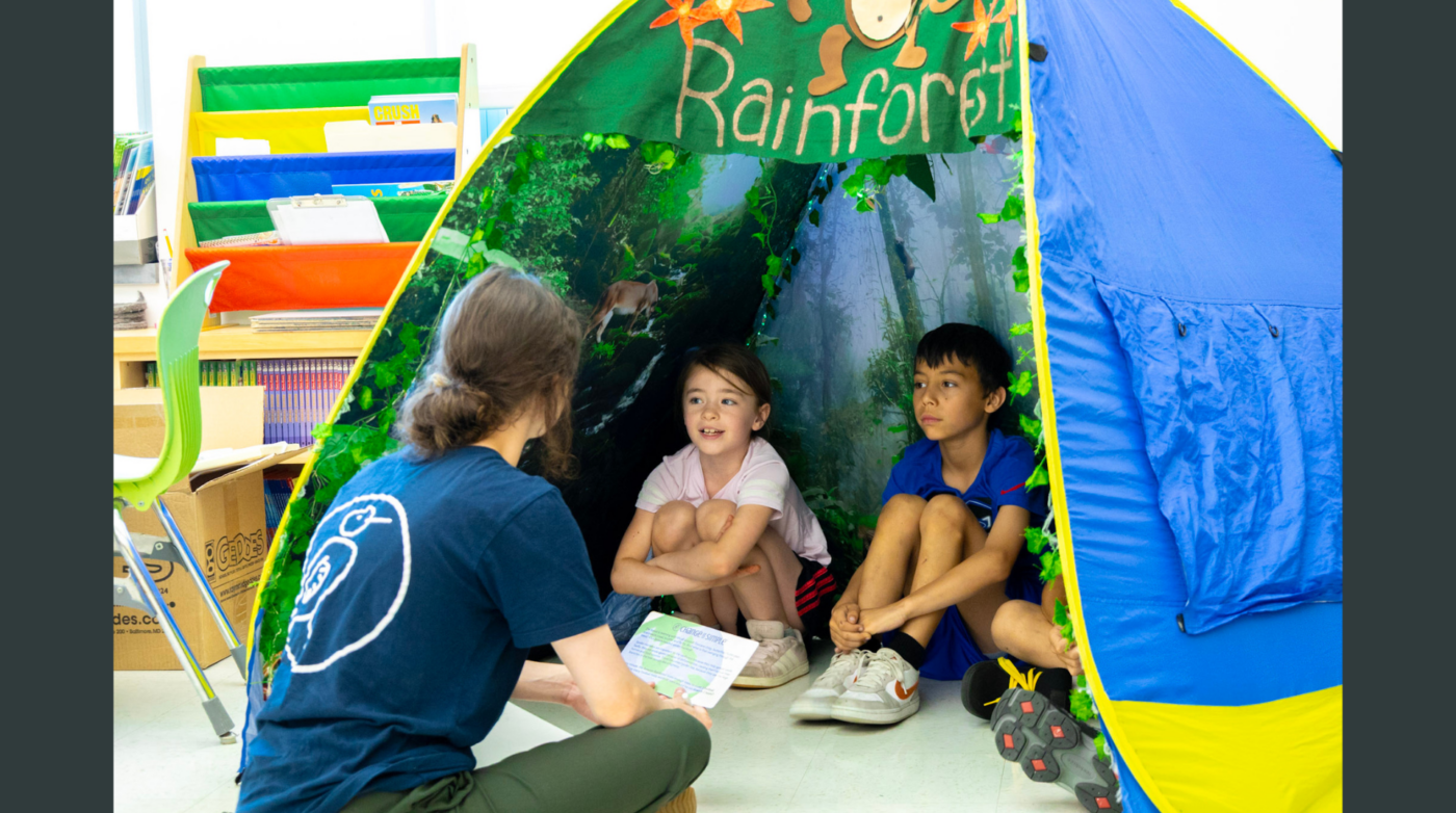 An adult sits on the floor, facing two children in a tent labeled "Rainforest" during a Change is Simple classroom activity. The children listen attentively, surrounded by shelves with colored bins, books, and a green chair in the background.