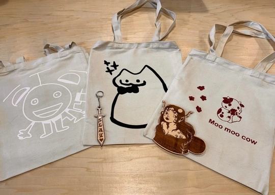 Three beige canvas tote bags from the Creativity Lab are displayed on a wooden surface, each featuring a unique cartoon design. Two keychain accessories—a sword-shaped one and one with a brown-haired character—add extra charm to the bags.
