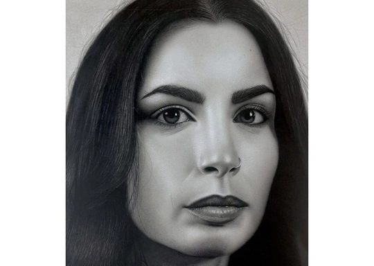 A black and white graphite portrait drawing of a person with long dark hair, clear skin, prominent eyebrows, and a small nose piercing. The individual is facing forward with a neutral expression against a plain, light-colored background.