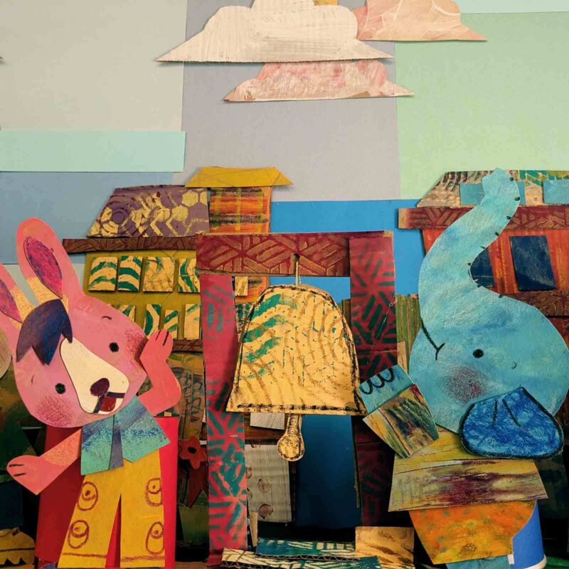 A pink rabbit and a blue elephant, made from colorful, textured paper, stand in front of a cardboard bell and a background of layered paper clouds and buildings. The rabbit waves while the elephant touches the bell.