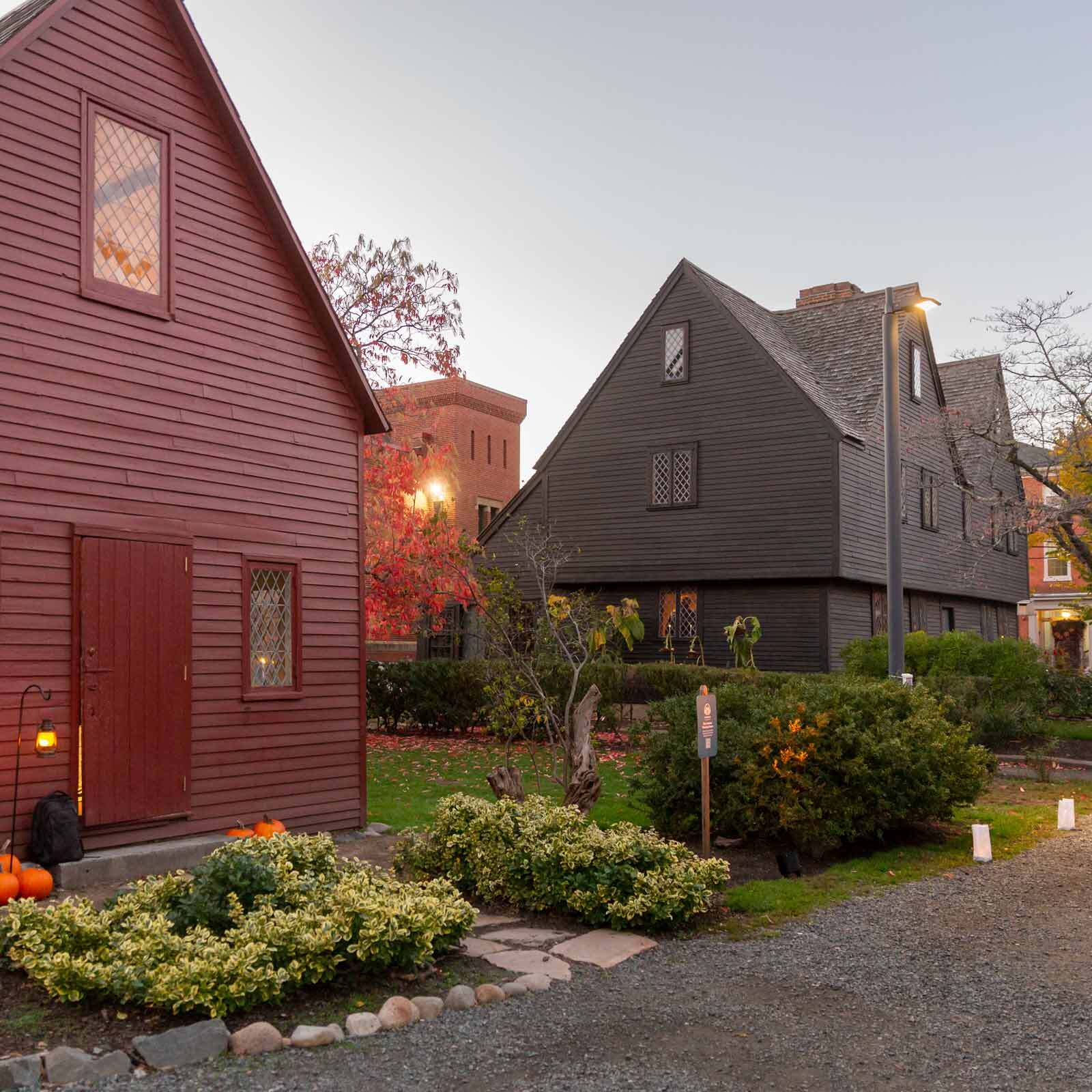 Two historic wooden houses with steep roofs, one red and one dark brown, stand along a gravel path surrounded by shrubs and pumpkins. Lanterns and paper luminaries line the path. Trees with autumn leaves are visible in the background.