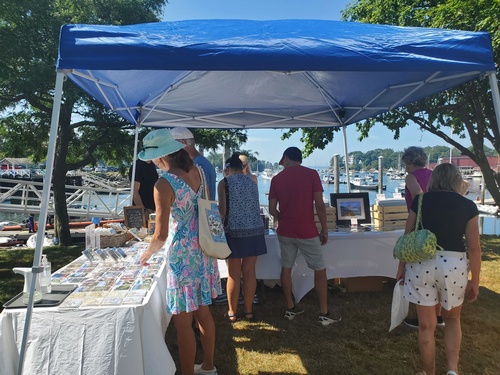 People browse items on tables under a blue canopy at an outdoor market near a marina. The sky is clear and boats are visible in the background. Some shoppers are wearing hats and casual summer clothing.