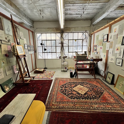 An art studio with large windows, exposed beams, and rugs on the floor. Home to Gallows Hill Artists, this space displays artworks on walls and easels. A desk with art supplies, a rolling chair, and framed pieces lend a creative air.