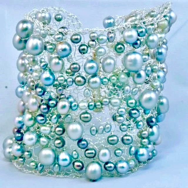 A cylindrical mesh structure features an irregular arrangement of variously sized, iridescent white and light blue pearls woven into metallic wire, set against a plain light background.
