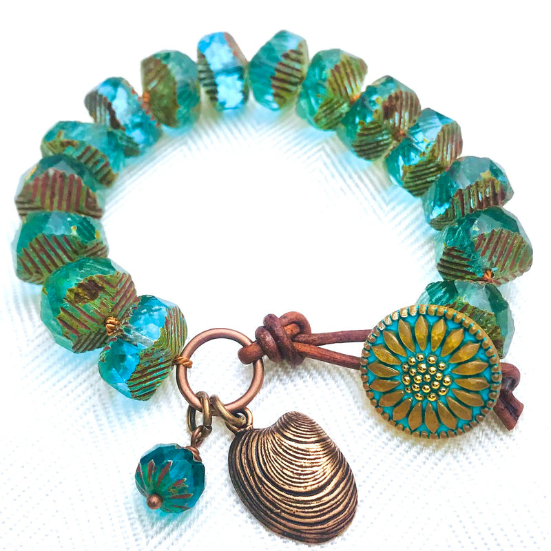 A bracelet with faceted blue-green beads and gold accents, a sunflower button clasp, a round charm with a sunflower design, a metallic shell charm, and a small matching bead charm. The bracelet is arranged in a circle on a white background.