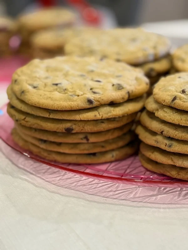 A close-up view of two neat stacks of large chocolate chip cookies on a pink glass plate, with more cookies blurred in the background. The cookies are golden brown and placed on a light-colored tablecloth.