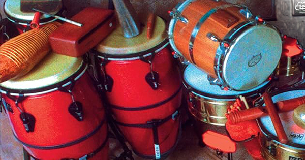 A collection of red conga drums, a snare drum, a güiro, and various percussion instruments are grouped together against a wall.