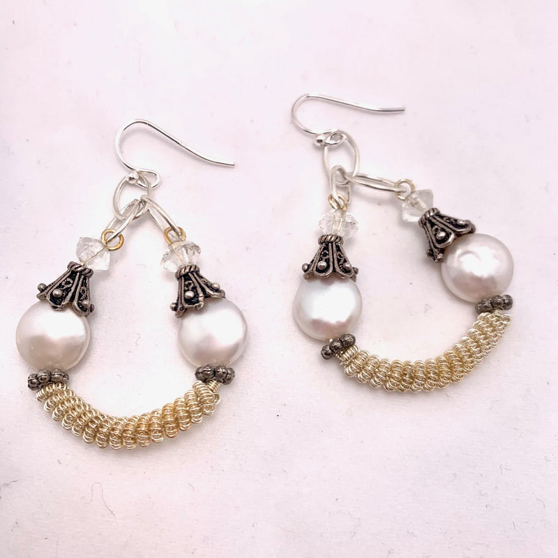 A pair of dangling earrings with large white pearls, ornate silver and black bead caps, small clear beads, and a curved section of coiled gold wire at the bottom, displayed on a white background.